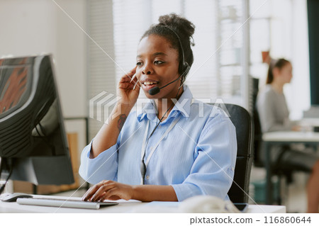 Young African American female worker dressed in pastel blue blouse helping client remotely via headphones while looking at details on monitor 116860644