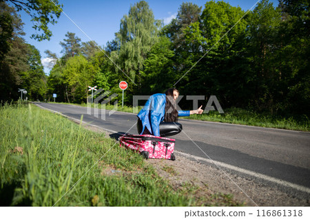 Bored of waiting to hitchhike, a teenager waits at the side of a road leading through a leafy forest on a sunny day. Bored of waiting to hitchhike, a teenager waits at the side of a road leading through a leafy forest on a sunny day. 116861318