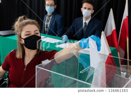 A woman in a wheelchair is trying to throw an envelope with a ballot paper into the ballot box. A woman in a wheelchair is trying to throw an envelope with a ballot paper into the ballot box. 116861383