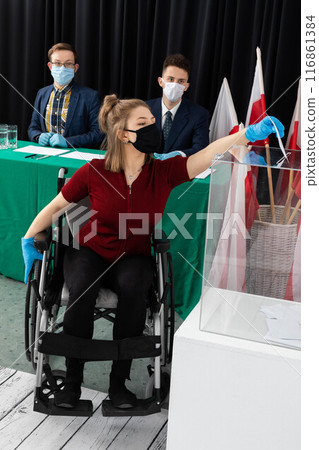 A woman in a wheelchair is trying to throw an envelope with a ballot paper into the ballot box. 116861384