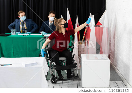 A woman in a wheelchair is trying to throw an envelope with a ballot paper into the ballot box. A woman in a wheelchair is trying to throw an envelope with a ballot paper into the ballot box. 116861385