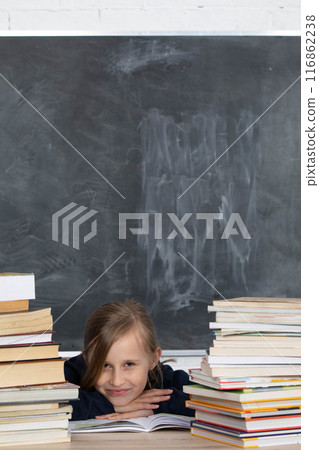 Girl sits in classroom among heap of school lector. Satisfied schoolgirl. School board in the background. Girl sits in classroom among heap of school lector. Satisfied schoolgirl. School board in the background. 116862238