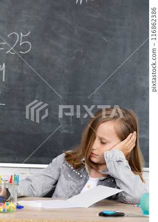 The schoolgirl sits at a school bench and supports her head with her hands in anticipation of a teacher. 116862386