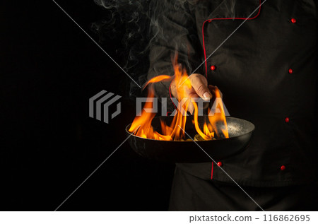The cook cooking food on a burning frying pan with flame. Restaurant and hotel service concept on a black background with space for advertising. Flambe or cooking vegetables in natural fire conditions 116862695