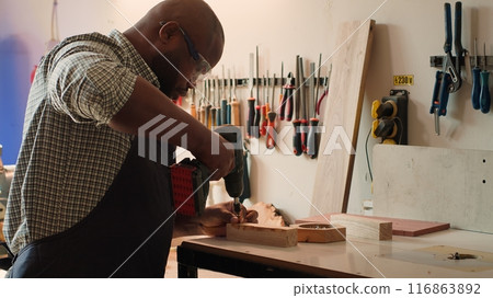 African american man in carpentry shop using power drill to create holes for dowels in wooden board. Craftsperson sinks screws into wood block with electric tool, doing precise drilling, camera A African american man in carpentry shop using power drill to create holes for dowels in wooden board. Craftsperson sinks screws into wood block with electric tool, doing precise drilling, camera A 116863892