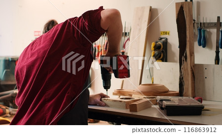 Woodworker in assembly shop using power drill to create holes for dowels in wooden board. Carpenter sinking screws into wooden surfaces with electric tool, doing precise drilling for seamless joinery 116863912