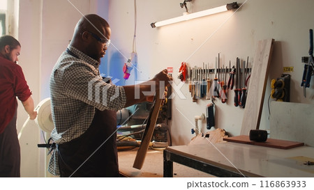 BIPOC carpenter in woodworking shop inspecting lumber piece before assembling furniture, checking for scratches. Cabinetmaker evaluates timber block, picking right tool from rack for it, camera B BIPOC carpenter in woodworking shop inspecting lumber piece before assembling furniture, checking for scratches. Cabinetmaker evaluates timber block, picking right tool from rack for it, camera B 116863933