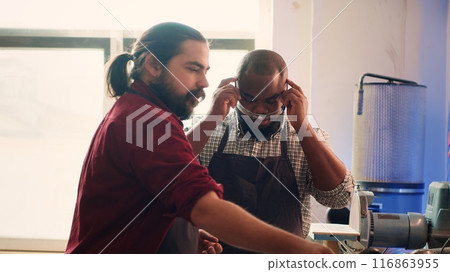 Master in carpentry shop instructing BIPOC worker to wear protection gear before assembling furniture. Cabinetmaker making sure apprentice uses safety earmuffs and glasses before starting work, camera 116863955