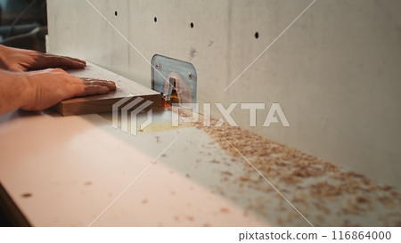 Carpenter in studio puts wood block through spindle moulder, creating smooth edges on wooden pieces. Cabinetmaker in joinery using heavy machinery to craft joints for furniture, camera B close up 116864000