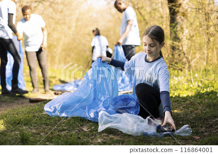 Young girl activist collecting trash and recycling in disposal bags, clearing the natural forest environment and combating illegal dumping. Ecologic volunteer grabbing plastic waste and junk. Young girl activist collecting trash and recycling in disposal bags, clearing the natural forest environment and combating illegal dumping. Ecologic volunteer grabbing plastic waste and junk. 116864001