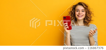 Cheerful woman in striped shirt holding red smartphone against yellow background, portraying modern communication and happiness. 116867376