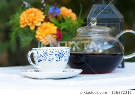 Patio table with cup of tea, pot, flowers and lantern. 116872311