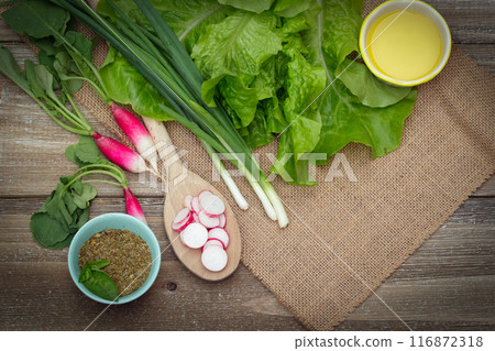 Fresh ripe vegetables on the wooden table with burlap table cloth - raddish sliced on a big spoon and whole, green onion, lettuce, dried herbs in a little prep bowl with basil leaves and olive oil are 116872318