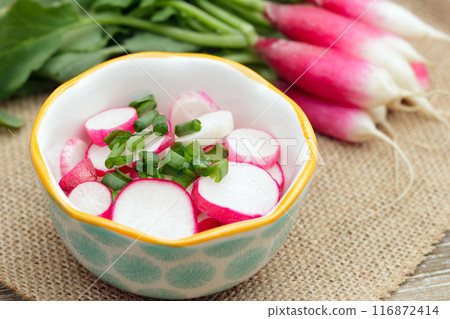 Salad of radishes and green onion in a little bowl on the burlap cloth on a wooden table and a bunch of French breakfast radishes behind it. 116872414