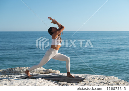 A woman in white sports clothes practices yoga exercises outdoors among white cliffs overlooking the sea 116873843