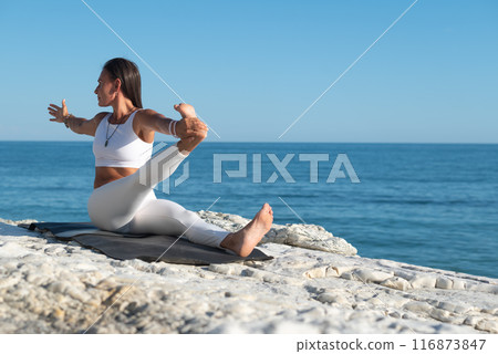A woman in white sports clothes practices yoga exercises outdoors among white cliffs overlooking the sea 116873847