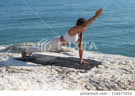 A woman in white sports clothes practices yoga exercises outdoors among white cliffs overlooking the sea 116873858