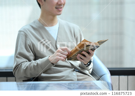Smiling young man relaxing on chair with book and looking out of window at coffee shop 116875723