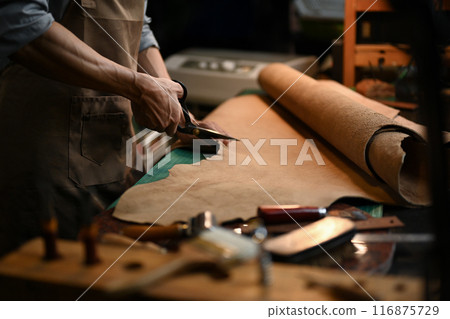Cropped shot of a craftsman cutting a large piece of leather with precision scissors in his workshop Cropped shot of a craftsman cutting a large piece of leather with precision scissors in his workshop 116875729