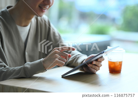 Smiling young Asian man sitting near window at coffee shop and reading book Smiling young Asian man sitting near window at coffee shop and reading book 116875757