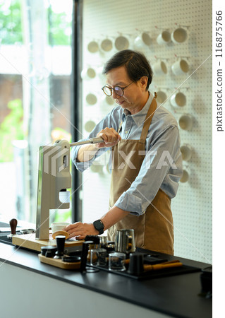 Senior male barista wearing an apron preparing a fresh cup of coffee using a modern espresso machine Senior male barista wearing an apron preparing a fresh cup of coffee using a modern espresso machine 116875766