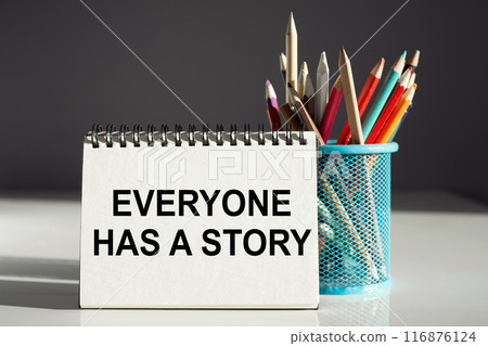 A white notebook with the inscription EVERYONE HAS A STORY standing near a blue pencil holder on white and gray background. Business concept 116876124