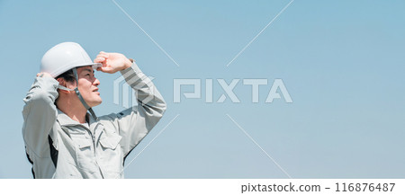 An Asian male worker in work clothes wearing a helmet and looking up at the sky under a blue sky 116876487