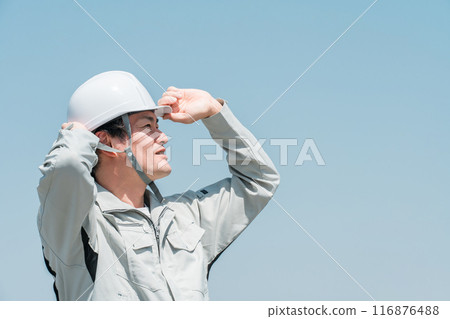 一名身穿工作服的亞洲男工戴著頭盔,仰望藍天下的天空 一名身穿工作服的亞洲男工戴著頭盔,仰望藍天下的天空 116876488