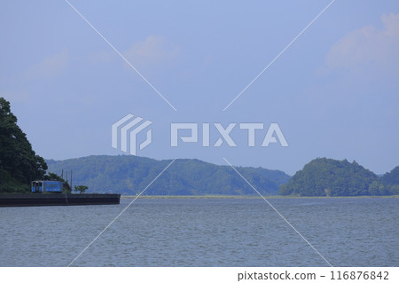 A local train with a blue-bodied carriage on the Hanasaki Line running along Lake Akkeshi in Hokkaido on a calm, sunny summer day 116876842