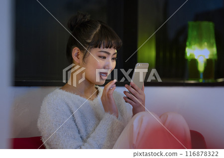 Smiling young woman operating a smartphone at home at night image Smiling young woman operating a smartphone at home at night image 116878322