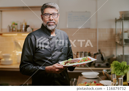 Proud middle aged chef dressed in black uniform smiling widely while demonstrating his served dish Proud middle aged chef dressed in black uniform smiling widely while demonstrating his served dish 116879732