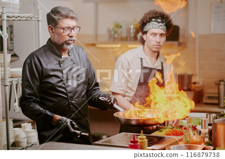 Middle aged chef cooking meat with fire while his best cook watching process Middle aged chef cooking meat with fire while his best cook watching process 116879738