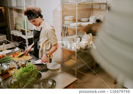 High angle shot of curly-haired young cook reading recipe while chopping vegetables on wooden cutting board 116879748