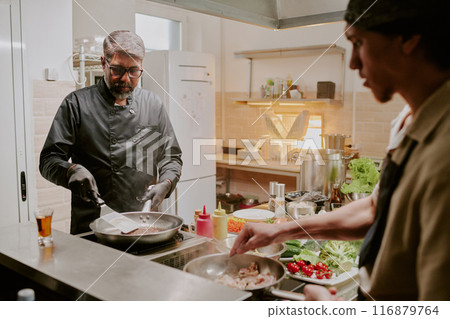 Professional Indian chef dressed in black jacket and wearing black latex gloves frying black angus steak while his sous chef frying bacon Professional Indian chef dressed in black jacket and wearing black latex gloves frying black angus steak while his sous chef frying bacon 116879764