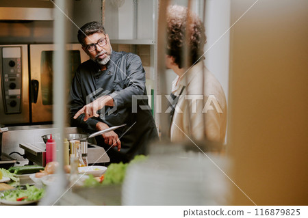 Middle aged chef with gray beard giving advices to his sous chef while he frying ingredients Middle aged chef with gray beard giving advices to his sous chef while he frying ingredients 116879825