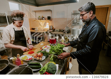 Middle aged chef and his young cook preparing food for guests in modern kitchen of luxury restaurant Middle aged chef and his young cook preparing food for guests in modern kitchen of luxury restaurant 116879828