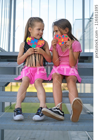 Two girls are sitting on the stairs holding heart-shaped postcards with puzzle pieces Two girls are sitting on the stairs holding heart-shaped postcards with puzzle pieces 116881895