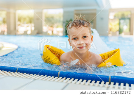 Portrait cute adorable little caucasian blond boy kid wear inflatable armband sit poolside indoor pool edge before swimming hotel spa thermal resort. Happy child enjoy summer vacation travel family Portrait cute adorable little caucasian blond boy kid wear inflatable armband sit poolside indoor pool edge before swimming hotel spa thermal resort. Happy child enjoy summer vacation travel family 116882172