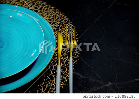 A view of half of the blue plates are standing one on one on a gold patterned napkin, gold cutlery is lying on the side: a fork and a knife on a black background with a place for text. The concept of 116882185