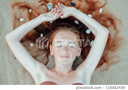 A woman peacefully lies on a serene beach, with stones in her hair, embodying relaxation and tranquility amidst the beauty of nature. Her expression reflects pure bliss and calmness 116882489