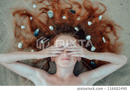 A woman with red hair covered in pebbles is seen on the beach, her hands over her eyes, embodying relaxation and inner peace. This scene captures a sense of tranquility and harmony with nature A woman with red hair covered in pebbles is seen on the beach, her hands over her eyes, embodying relaxation and inner peace. This scene captures a sense of tranquility and harmony with nature 116882490