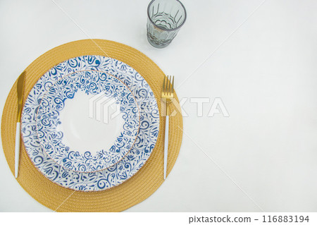 The plates are white with a blue pattern on the edges, they stand on a yellow straw napkin, a knife and fork are on the sides, a blue glass on a white background is visible behind them. The concept of 116883194