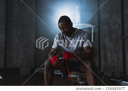 Fitness workout in gym. African American man fighter preparing for fight wrapping hands with red boxing wraps sports protective bandages in gym. Strong man ready for fight boxing sparring training Fitness workout in gym. African American man fighter preparing for fight wrapping hands with red boxing wraps sports protective bandages in gym. Strong man ready for fight boxing sparring training 116885977