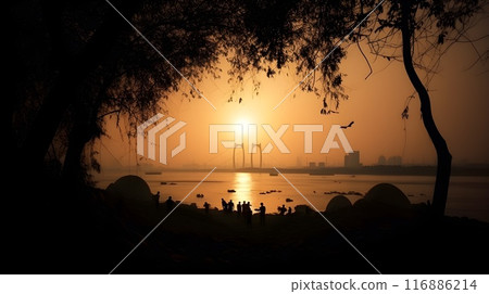 Tranquil beach at dawn: calm ocean, tree silhouette, golden sunset, bridge and city in the background. 116886214