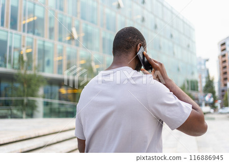 Back view African American man talking on smartphone outdoor. Unrecognizable person guy with cell phone walking on street in city. Man making answering call by cellphone having conversation by mobile Back view African American man talking on smartphone outdoor. Unrecognizable person guy with cell phone walking on street in city. Man making answering call by cellphone having conversation by mobile 116886945