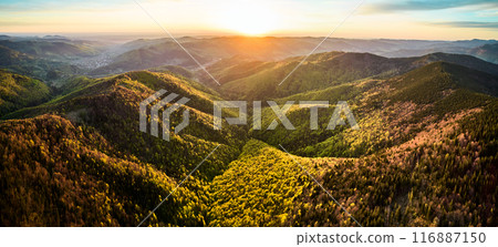Aerial view of breathtaking sunrise over rolling mountains covered in mix of lush green and autumnal foliage, with dramatic clouds streaking across sky, casting warm, golden light over the landscape. 116887150