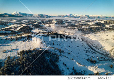 Aerial view of snow-covered valley with winding road passing through small village. Landscape features gently rolling hills, dense forests, and distant mountains under clear blue sky, with soft mist. 116887152