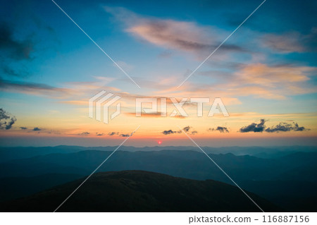 Sun sets behind rolling hills, casting warm, golden glow across sky. Wispy clouds, tinged with hues of orange and yellow, stretch across deepening blue sky. Serene and expansive landscape. Sun sets behind rolling hills, casting warm, golden glow across sky. Wispy clouds, tinged with hues of orange and yellow, stretch across deepening blue sky. Serene and expansive landscape. 116887156