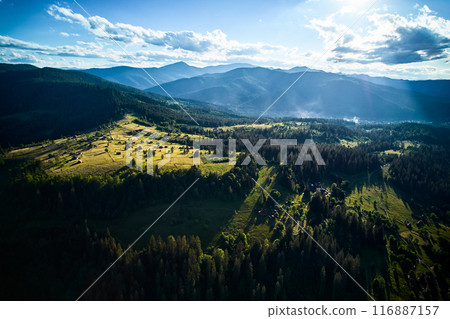 Aerial view of lush, green valley dotted with small houses and winding roads, surrounded by dense forests and rolling hills. Sunlight filters through scattered clouds. Mountains rise under blue sky. Aerial view of lush, green valley dotted with small houses and winding roads, surrounded by dense forests and rolling hills. Sunlight filters through scattered clouds. Mountains rise under blue sky. 116887157