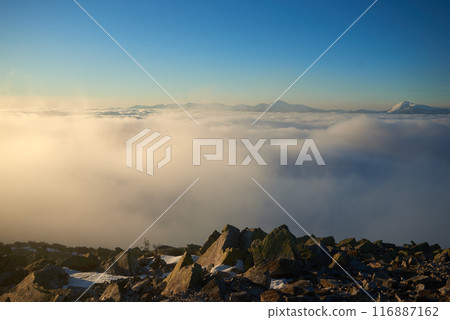 Sunrise over snow-covered mountain peak. Warm golden light of rising sun bathes clouds in soft hues of orange and pink. Snow and scattered rocks in foreground add texture and depth to scene. Sunrise over snow-covered mountain peak. Warm golden light of rising sun bathes clouds in soft hues of orange and pink. Snow and scattered rocks in foreground add texture and depth to scene. 116887162